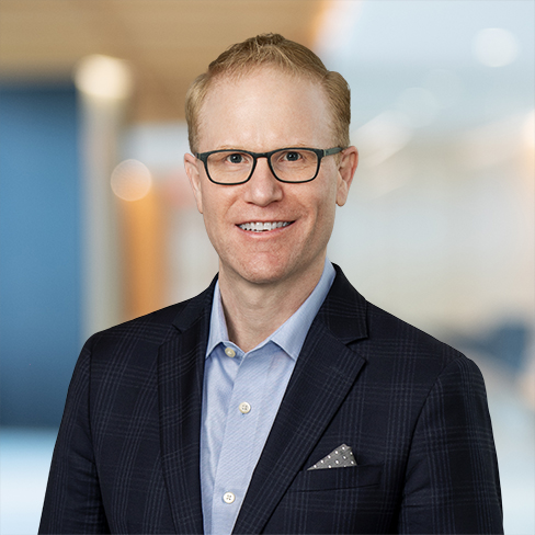 A man with short blond hair and glasses is wearing a navy blazer, light blue shirt, and pocket square, standing confidently in a modern corporate law office.