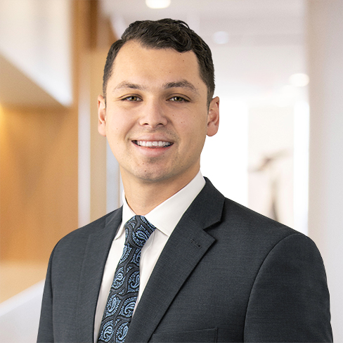 A young man in a dark suit, white shirt, and patterned tie stands indoors, smiling at the camera with a blurred corporate law office in the background.