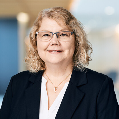 A middle-aged woman with curly blonde hair, glasses, and a dark blazer over a white blouse stands in a modern corporate law office.