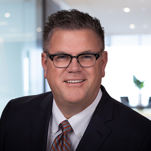 Man wearing glasses, a dark suit, white shirt, and striped tie, smiling in a modern law office setting with glass walls and bright lighting—an example of professional Chicago lawyers at work.