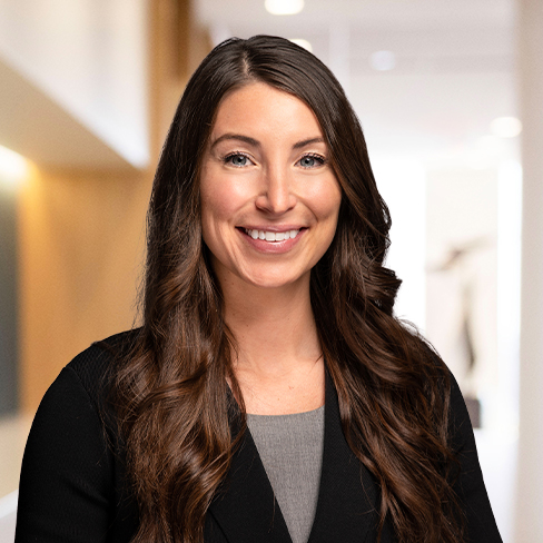 Woman with long brown hair wearing a gray top and black blazer, smiling, standing in a well-lit corporate law office hallway with a blurred background.