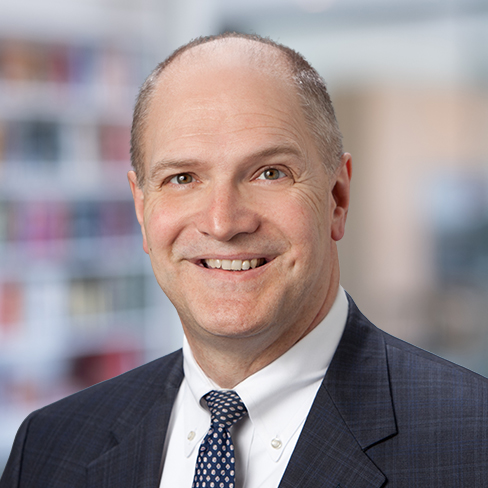 A middle-aged man in a suit and tie smiles at the camera in an indoor setting with blurred bookshelves, reflecting the professional ambiance of a corporate law office.