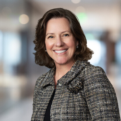 Woman with shoulder-length brown hair wearing a textured blazer, smiling, standing in a blurred corporate law office environment.