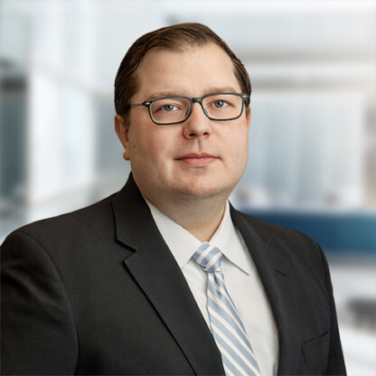 A man wearing glasses, a dark suit, a white shirt, and a light striped tie stands in front of a blurred office background, representing chicago lawyers specializing in intellectual property law.