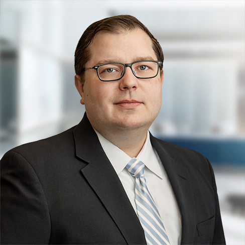 A man wearing glasses, a dark suit, a white shirt, and a light striped tie stands in front of a blurred office background, representing chicago lawyers specializing in intellectual property law.