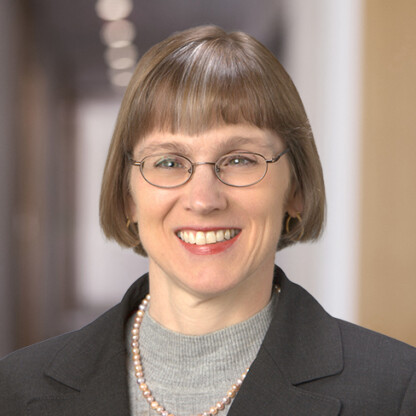 A woman with short light brown hair and glasses, wearing a gray turtleneck, dark blazer, and pearl necklace, smiles in a blurred corporate law office hallway background.