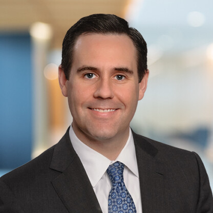 A man in a dark suit, white shirt, and patterned blue tie is smiling in an office setting with a blurred background, reflecting the professional environment common in law offices specializing in litigation support.