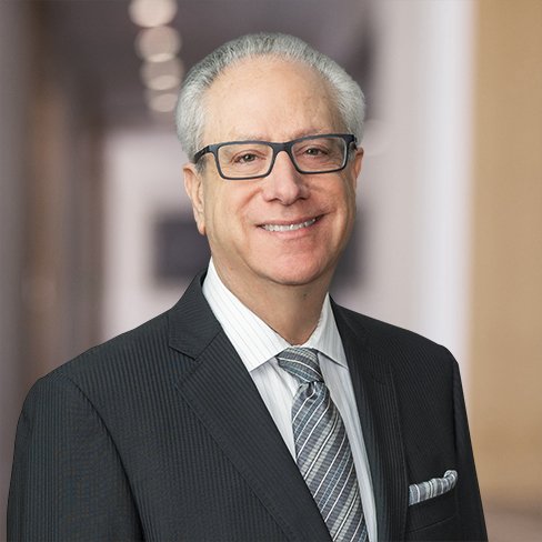 A man with gray hair and glasses, wearing a dark suit, striped shirt, and patterned tie, stands in a blurred hallway of corporate law offices.