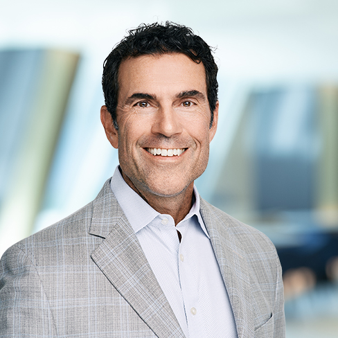 Smiling man with dark hair wearing a light gray plaid suit jacket and white shirt, standing in a modern, blurred corporate law office—a professional among Chicago lawyers.