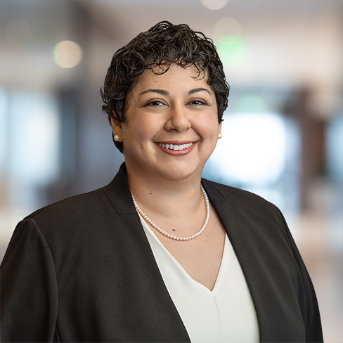 A woman with short curly hair, wearing a black blazer, white blouse, and pearl necklace, smiles while standing in a blurred law offices setting, reflecting professionalism in intellectual property law.