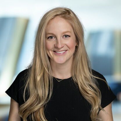 A woman with long blonde hair wearing a black top smiles at the camera in a modern law offices setting, with a blurred background.
