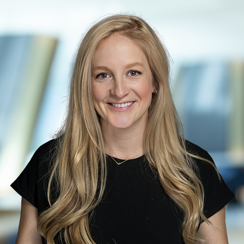 A woman with long blonde hair wearing a black top smiles at the camera in a modern law offices setting, with a blurred background.