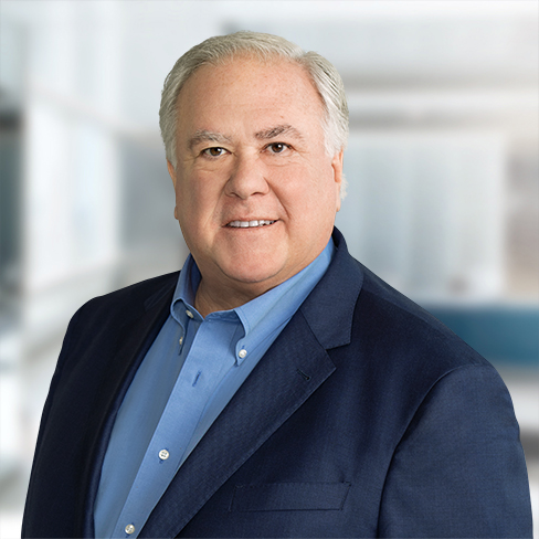 A middle-aged man with gray hair wearing a blue shirt and dark blazer, posing and smiling in a modern office setting, representing experienced lawyers in Chicago specializing in intellectual property law.