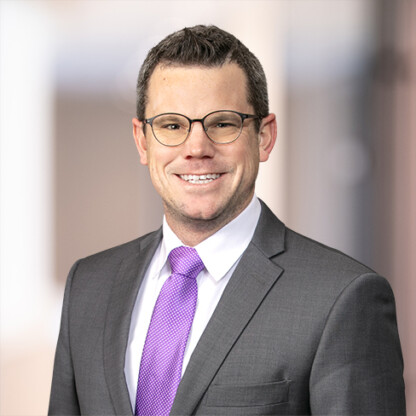 Man in a gray suit, white shirt, and purple tie, wearing glasses, smiling at the camera with a blurred indoor background—representing experienced lawyers in Chicago.