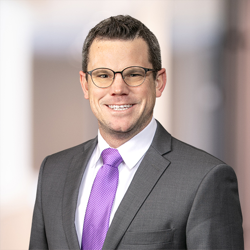 Man in a gray suit, white shirt, and purple tie, wearing glasses, smiling at the camera with a blurred indoor background—representing experienced lawyers in Chicago.