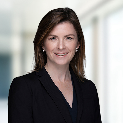 A woman with straight brown hair wearing a black blazer stands indoors against a blurred background, smiling at the camera, capturing the professional atmosphere of corporate law offices.