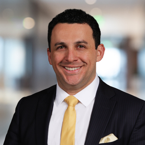 A man in a dark pinstripe suit, white shirt, and yellow tie smiles at the camera with a blurred law offices background, representing professional lawyers in Chicago.