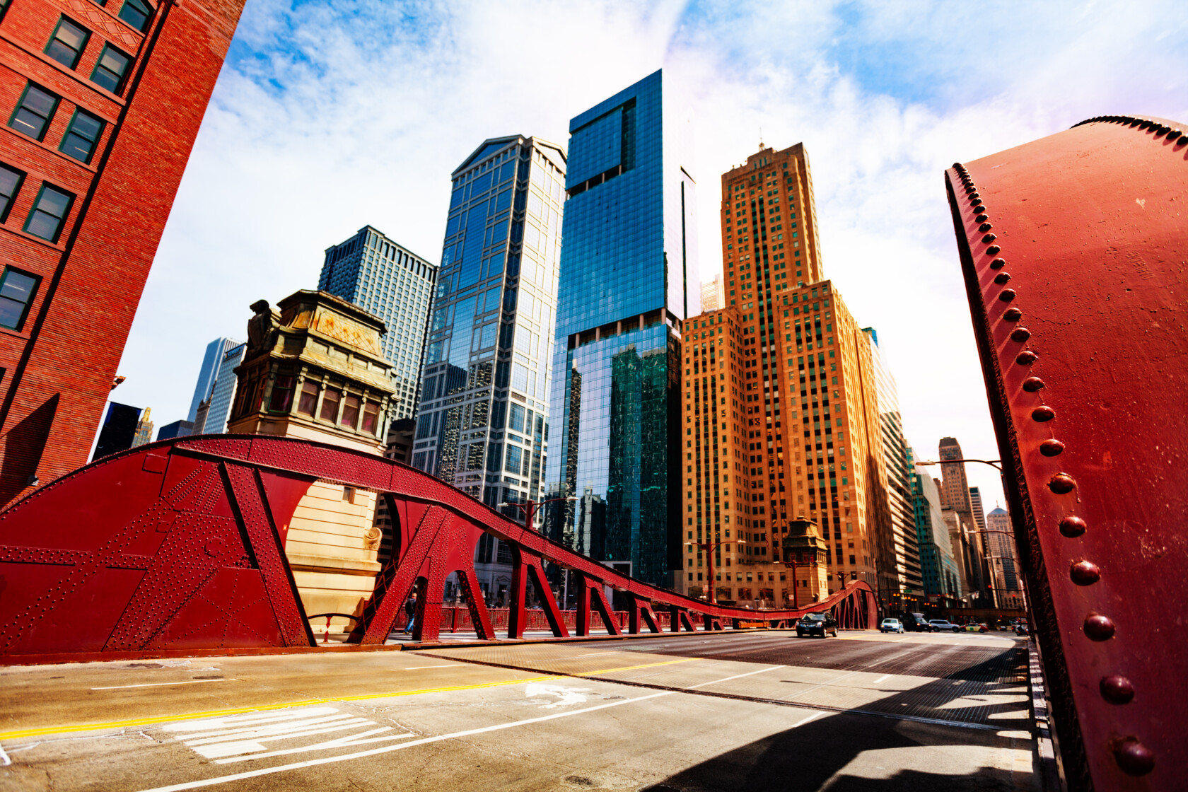 Red metal bridge crosses in front of tall modern and historic skyscrapers, including a prominent corporate law office, all set against a partly cloudy sky.