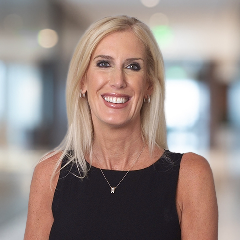 A woman with long blonde hair, wearing a sleeveless black top and a necklace, smiles while standing in a bright, blurred corporate law office setting.