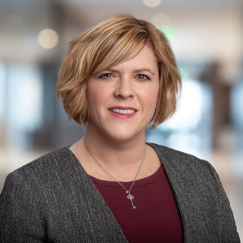 Woman with short blonde hair wearing a grey blazer and maroon top, standing in front of a blurred corporate law office background, smiling at the camera.