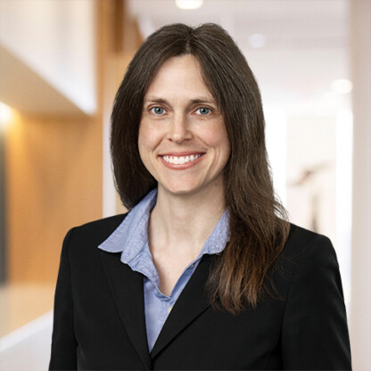 A woman with long brown hair wearing a black blazer and blue collared shirt stands smiling in a modern law office hallway, reflecting the professionalism of Chicago lawyers.
