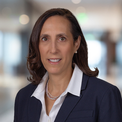 Woman with shoulder-length brown hair, wearing a navy blazer, white blouse, and pearl necklace, posed in a blurred corporate law office—reflecting the professionalism of lawyers in Chicago.