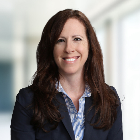 Woman with long brown hair wearing a navy blazer and striped shirt, smiling at the camera in a bright, blurred corporate law office setting.