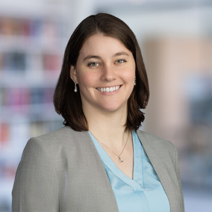 A woman with shoulder-length brown hair, wearing a gray blazer over a light blue top, stands smiling in front of a blurred office background at one of Chicago’s leading law offices specializing in intellectual property law.