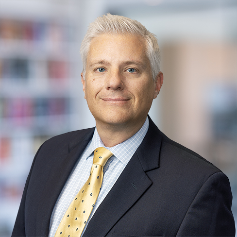 A man with short gray hair wearing a dark suit, light blue checkered shirt, and yellow tie stands confidently in a corporate law office environment.