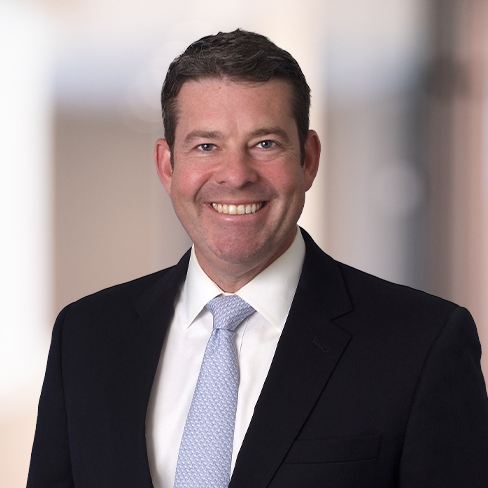 A man in a dark suit, white shirt, and light-colored tie smiles at the camera in a professional, blurred corporate law office setting.