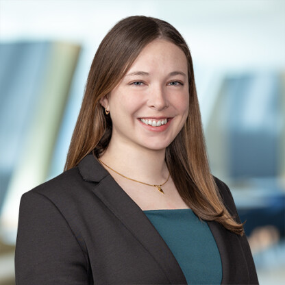 A woman with straight brown hair, wearing a dark blazer and teal top, smiles at the camera against a blurred office background, reflecting her professionalism in litigation support for law offices.