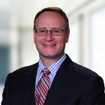A man wearing glasses, a dark suit, a striped shirt, and a multicolored striped tie poses in front of a blurred corporate law office, reflecting the professionalism of lawyers in Chicago.