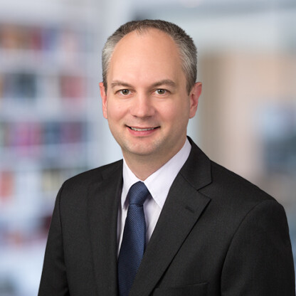 A man in a dark suit, white shirt, and blue tie smiles at the camera against a blurred corporate law office background.