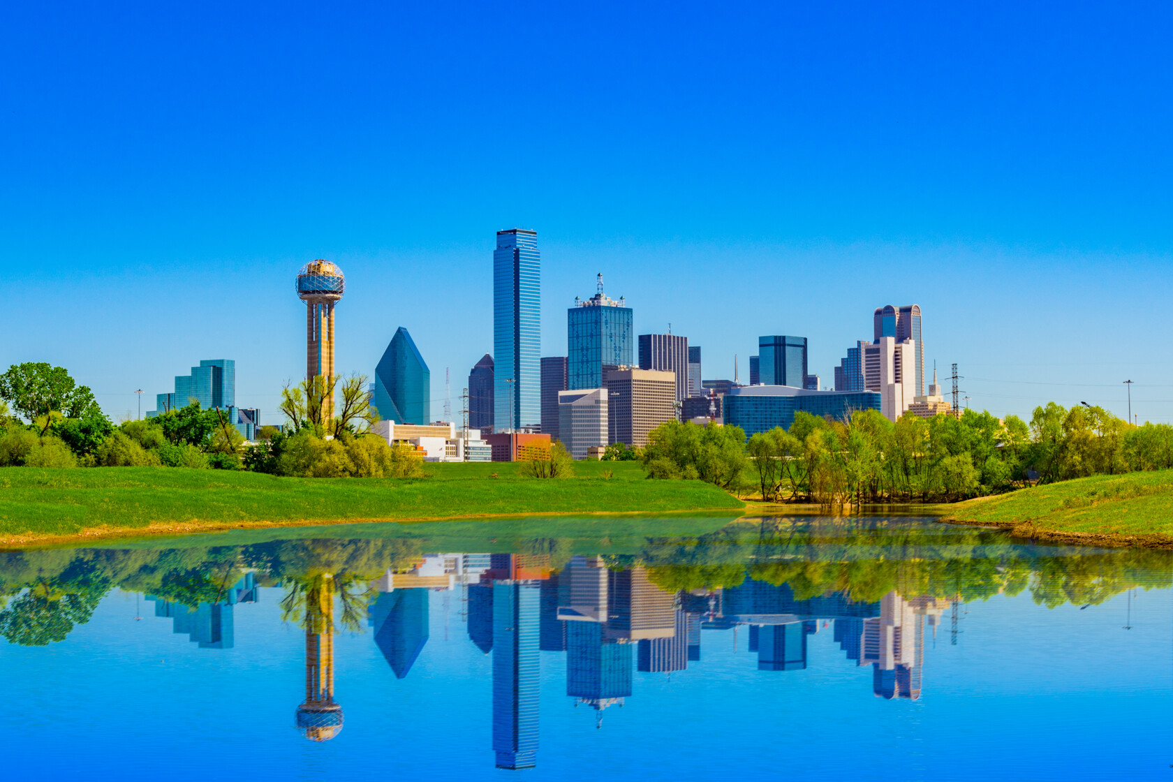 Dallas city skyline reflected in a calm body of water on a clear, sunny day, with green trees and grass in the foreground—an inspiring view for law offices offering litigation support.