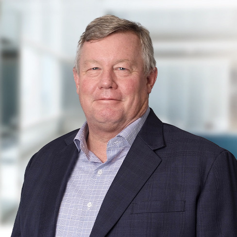 Middle-aged man with short gray hair wearing a dark blazer and checked shirt, standing in a bright, modern office setting specializing in litigation support and intellectual property law.