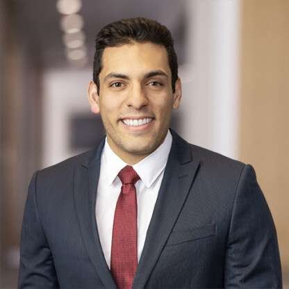 A man in a dark suit, white dress shirt, and red tie is smiling while standing in a modern corporate law office hallway.