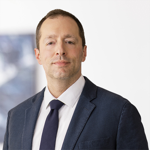 A man wearing a dark suit, white shirt, and dark tie poses against a light, blurred background, embodying the professionalism often found in law offices and litigation support settings.