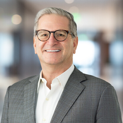 A man wearing glasses, a gray suit jacket, and a white shirt smiles in front of a blurred corporate law office background.