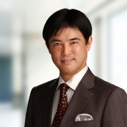 A man in a dark suit, white shirt, and patterned tie stands in front of a blurred law offices background, looking at the camera and smiling slightly.