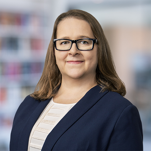 A woman with long brown hair and glasses, wearing a navy blazer and white top, stands in front of a blurred office or library background—reflecting the professionalism often seen among Chicago lawyers specializing in intellectual property law.