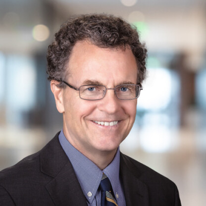 Man with short curly hair, glasses, and a dark suit smiling in an indoor office setting at one of the top law offices specializing in intellectual property law, with a blurred background.