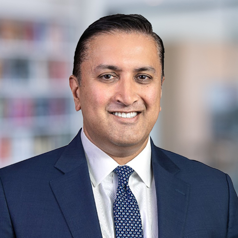 A man in a navy blue suit, white shirt, and patterned tie smiles at the camera with a blurred corporate law office background.