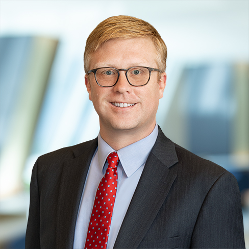 A man with short blond hair, glasses, and a suit with a red polka dot tie stands in front of a blurred law offices background, reflecting the professionalism of Chicago lawyers.