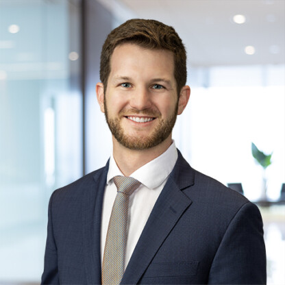 A man in a navy suit and striped tie stands in a modern law office with glass walls, smiling at the camera, embodying professionalism in intellectual property law.
