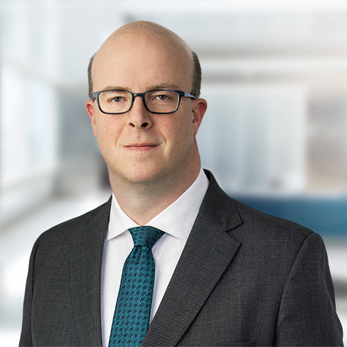 A man in a suit and tie with glasses stands in a corporate law office, looking at the camera with a neutral expression.