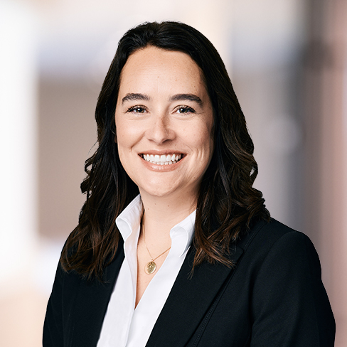 Woman with long brown hair wearing a black blazer and white shirt, smiling, in front of a blurred indoor background—representing experienced lawyers in Chicago.