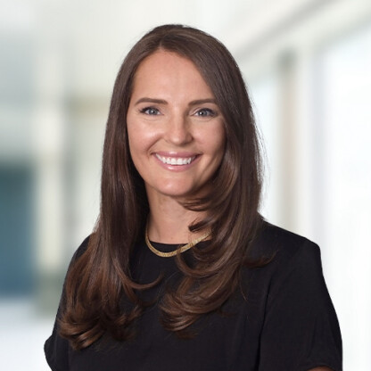 Woman with long brown hair wearing a black top and gold necklace, smiling, posed in front of a blurred corporate law office background.