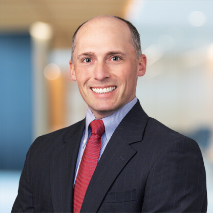 A man in a dark suit, light blue shirt, and red tie smiles at the camera in a modern corporate law office setting.