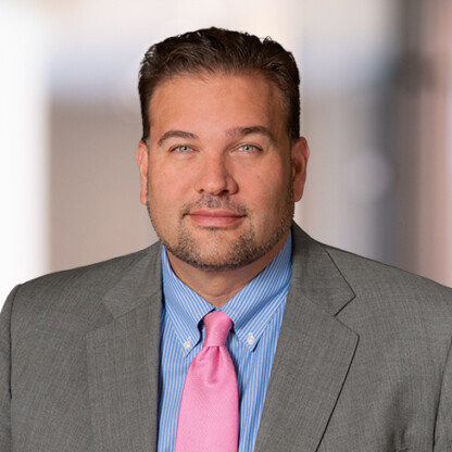 A man wearing a gray suit jacket, blue striped shirt, and pink tie poses for a professional headshot against a blurred indoor background, reflecting the polished image common in top Chicago lawyers’ law offices.