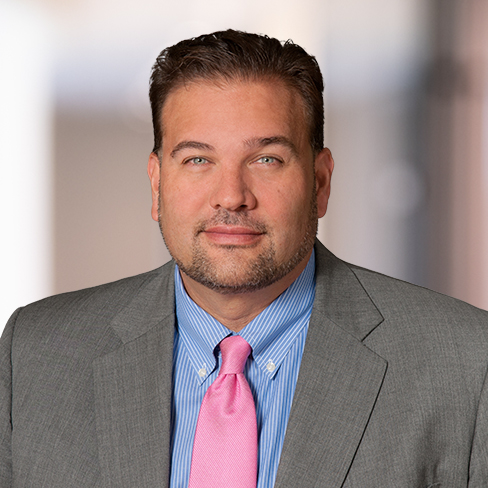 A man wearing a gray suit jacket, blue striped shirt, and pink tie poses for a professional headshot against a blurred indoor background, reflecting the polished image common in top Chicago lawyers’ law offices.
